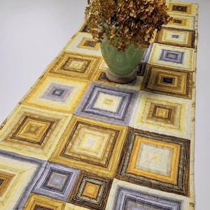 May include: A quilted table runner with a geometric pattern of squares in shades of yellow, brown, and purple. The runner is made of fabric and has a subtle sheen.
