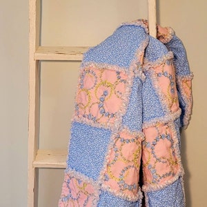 May include: A blue and pink patchwork quilt with a ruffled edge. The quilt is hanging on a white wooden ladder.