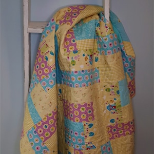 May include: A yellow, pink and blue patchwork quilt with a whimsical design hangs on a white wooden ladder.