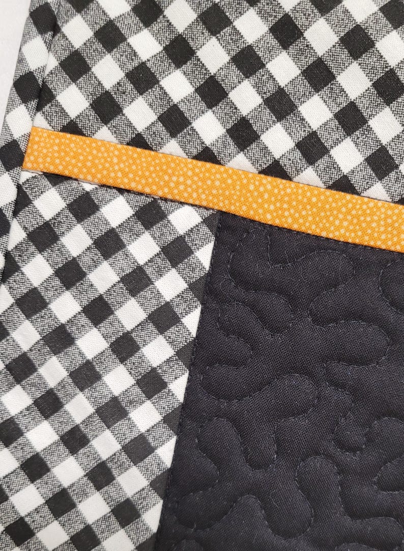 May include: A black and white checkered fabric with a bright orange trim. The fabric is sewn together with a black fabric with a quilted pattern.