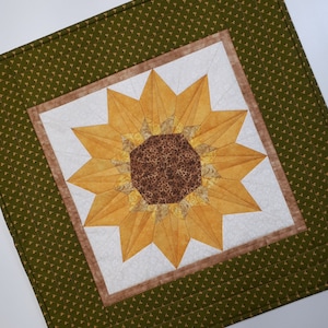 May include: A quilted wall hanging featuring a sunflower design with a brown center and yellow petals. The sunflower is surrounded by a brown and white frame and a green background with a small white pattern.