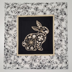 Peut inclure: Un élément décoratif mural carré et matelassé avec une bordure à motifs floraux noirs et blancs. Le centre présente un carré noir avec un motif de lapin brodé détaillé en blanc et or. Le lapin est rempli de motifs floraux.