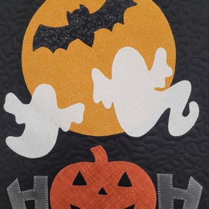 May include: A black quilted fabric with a white ghost, a black bat flying in front of a yellow moon, and an orange pumpkin with a carved face. The words "HOH" are on either side of the pumpkin.