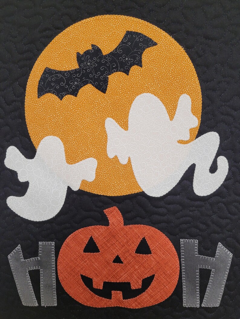 May include: A black quilted fabric with a white ghost, a black bat flying in front of a yellow moon, and an orange pumpkin with a carved face. The words "HOH" are on either side of the pumpkin.