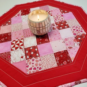 May include: A red octagonal table runner with a white and pink patchwork design featuring hearts and other patterns. A glass candle holder with a lit candle sits in the centre of the runner.