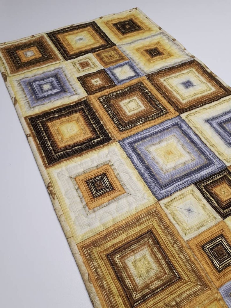 May include: A quilted table runner with a geometric pattern of squares in shades of brown, beige, and light blue.