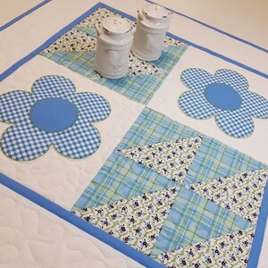 May include: A white quilted table runner with a blue border. The runner features a blue and white gingham flower design in each corner. Two white ceramic salt and pepper shakers are placed in the centre of the runner.