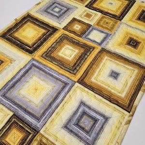 May include: A quilted table runner with a geometric pattern of squares in shades of brown, yellow, and gray. The squares are layered and have a concentric design.