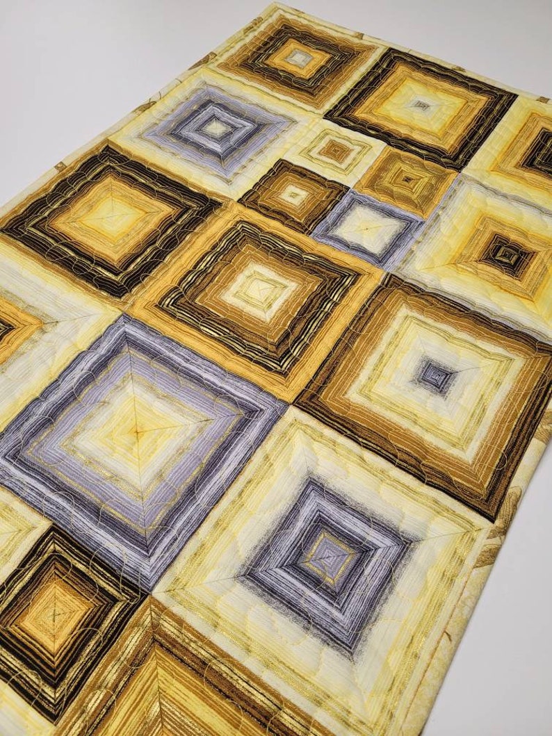 May include: A quilted table runner with a geometric pattern of squares in shades of brown, yellow, and gray. The squares are layered and have a concentric design.