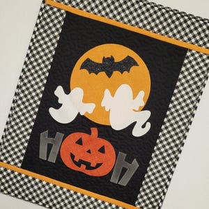 May include: A black and white checkered fabric with a black panel featuring a yellow moon with a black bat, two white ghosts, a pumpkin with a jack-o-lantern face, and two gray fence posts with the word "BOO" in gray letters.