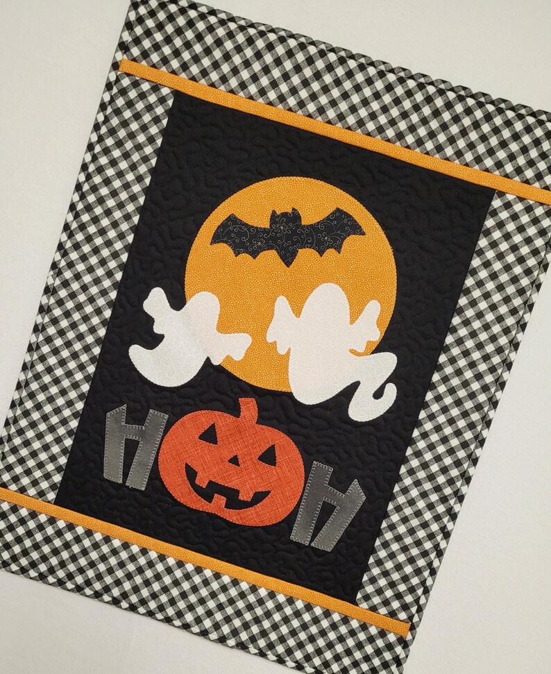 May include: A black and white checkered fabric with a black panel featuring a yellow moon with a black bat, two white ghosts, a pumpkin with a jack-o-lantern face, and two gray fence posts with the word "BOO" in gray letters.