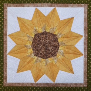 May include: A quilted wall hanging featuring a large, stylized sunflower with brown and yellow fabric. The sunflower is surrounded by a white background with a subtle floral pattern.