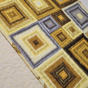 May include: A quilted table runner with a geometric pattern of squares in shades of yellow, brown, and gray. The runner has a cream-colored backing.