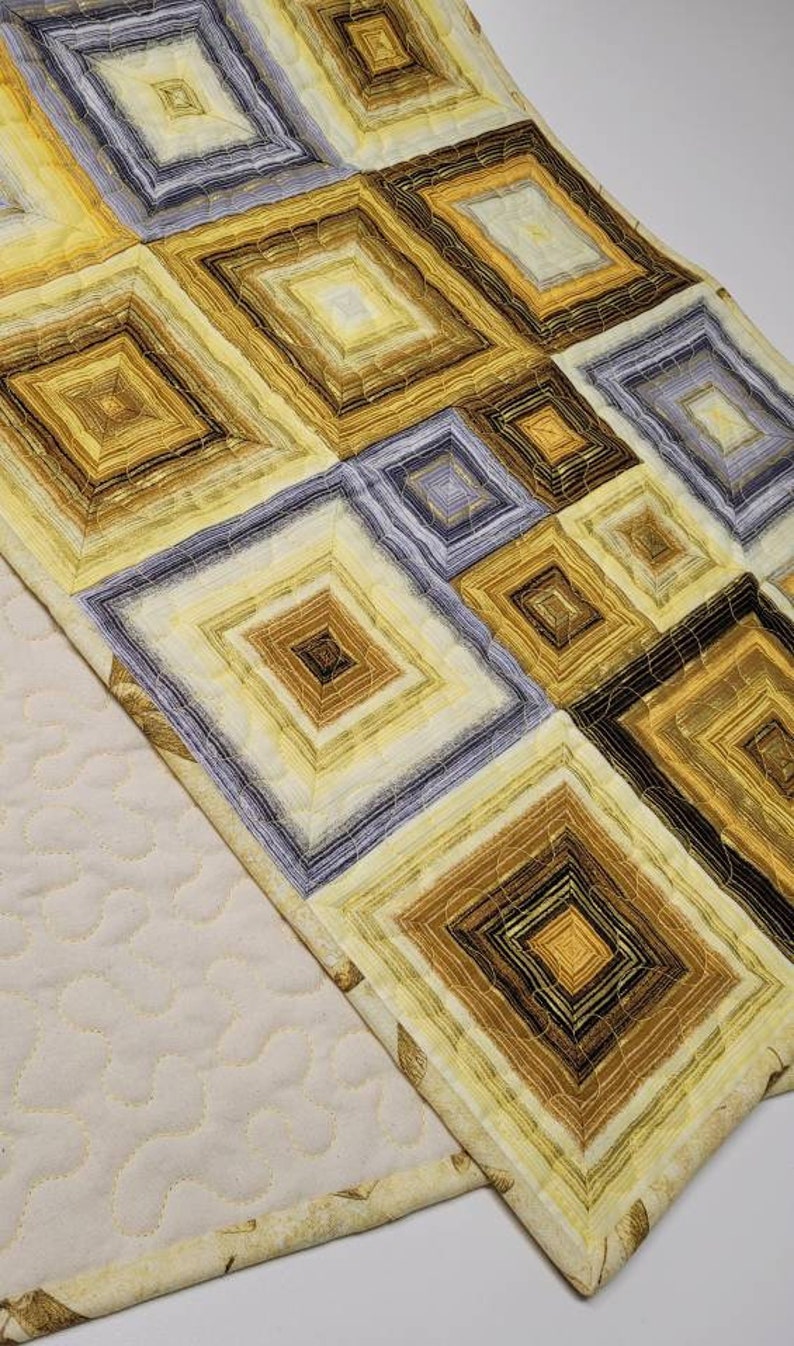 May include: A quilted table runner with a geometric pattern of squares in shades of yellow, brown, and gray. The runner has a cream-colored backing.