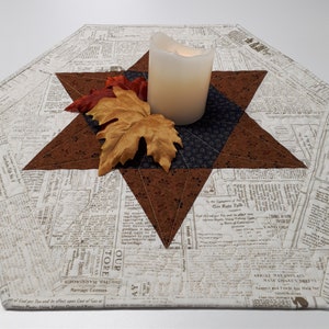 May include: A brown and white table runner with a star design. The star is made of brown fabric with a white outline. The table runner is made of a newspaper print fabric. A white candle sits in the centre of the star.