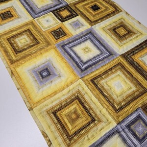May include: A table runner with a geometric pattern of squares in shades of yellow, brown, and gray. The squares are layered and quilted, creating a textured effect.