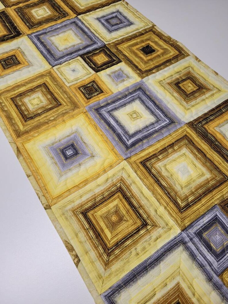 May include: A table runner with a geometric pattern of squares in shades of yellow, brown, and gray. The squares are layered and quilted, creating a textured effect.