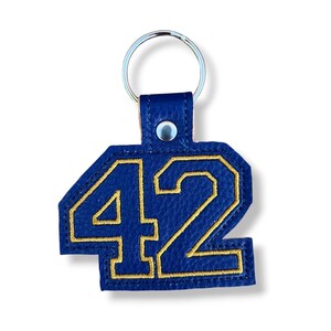 Op de afbeelding: Een blauwe sleutelhanger met het nummer 42 in gouden stiksels. De sleutelhanger is gemaakt van een leerachtig materiaal en heeft een zilveren sleutelring. Het nummer is in een vetgedrukte lettertype.