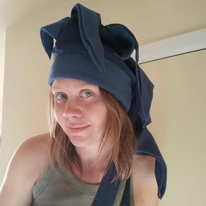 May include: A dark blue fleece hat with long, pointed flaps. The hat is worn by a person wearing a green ribbed tank top. The hat's flaps extend down the sides and back, creating a unique, whimsical design.