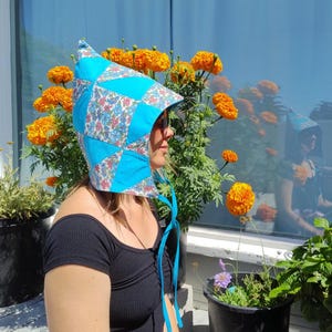 May include: A blue and floral patchwork bonnet with a pointed top and ties. The bonnet features a mix of solid blue and patterned fabric with a floral design. The bonnet is worn outdoors, with orange flowers in the background.