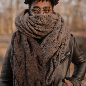 Puede incluir: Una persona lleva una bufanda de punto marrón envuelta alrededor de su cuello y una chaqueta de cuero negro. La bufanda es gruesa y tiene un patrón de punto de cable. La persona lleva gafas redondas y tiene el pelo corto y rizado.