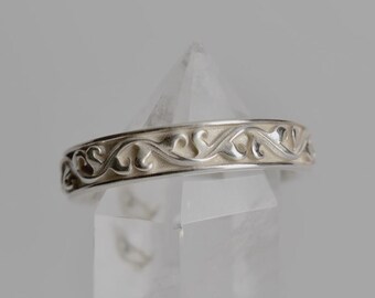 May include: A silver-toned metal ring featuring a continuous embossed scrollwork pattern along the outer band. The ring is positioned horizontally around the pointed tip of a clear, faceted, transparent quartz crystal point.