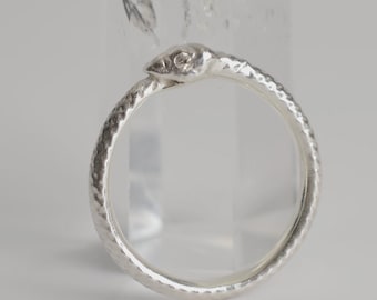 May include: A silver ring shaped like a coiled snake with textured scales along the band and a detailed head. The ring is positioned in front of a clear, pointed quartz crystal, showcasing the metallic finish and the intricate serpent design.