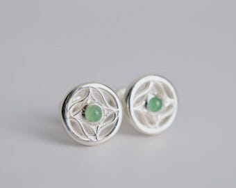 May include: A pair of circular silver stud earrings featuring an openwork lattice design. Each earring is centered with a small, smooth, round light green gemstone. The metal has a polished, reflective finish, and the earrings are presented as a set.