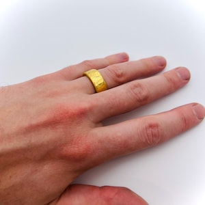May include: A gold ring with a textured surface. The ring is worn on the finger of a person's hand.