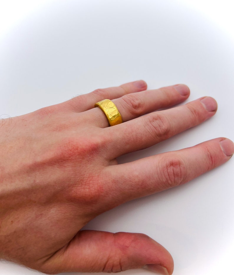 May include: A gold ring with a textured surface. The ring is worn on the finger of a person's hand.