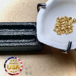 May include: A white ceramic crucible with a metal handle, filled with a pile of small, shiny gold nuggets. The crucible is sitting on a black, textured surface.  Made in USA.