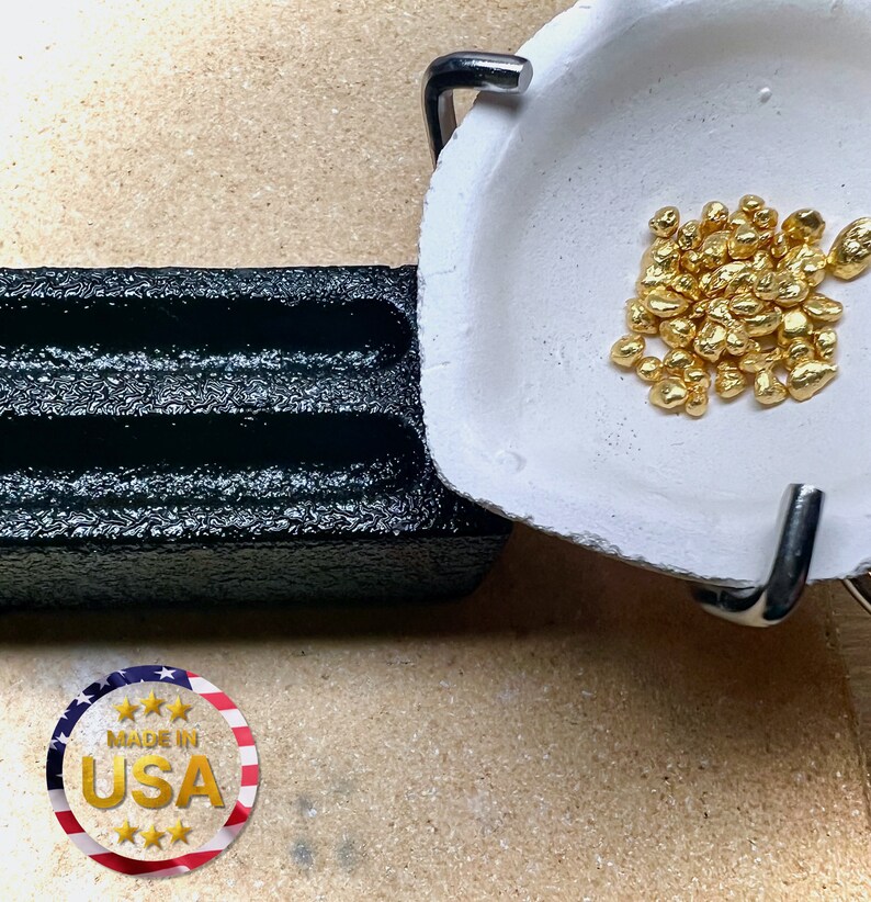 May include: A white ceramic crucible with a metal handle, filled with a pile of small, shiny gold nuggets. The crucible is sitting on a black, textured surface.  Made in USA.