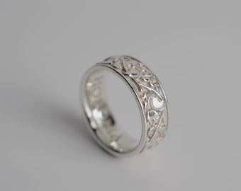May include: A silver ring featuring an intricate, raised floral or vine-like relief pattern covering the outer band. The metal has a polished, reflective finish, and the interior of the band appears smooth with visible engraved text along the inner edge.