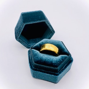 May include: A gold ring with a textured surface sits inside a teal velvet ring box.