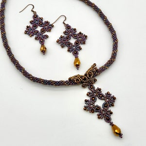May include: A beaded necklace and earrings set with a purple and gold color scheme. The necklace features a pendant with a butterfly design and a dangling flower-shaped charm. The earrings are also flower-shaped.