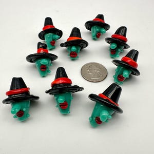 May include: A collection of green glass witch head beads. Each bead features a black hat with a red band, red lips, and a small nose. The beads are arranged on a white surface, with a quarter coin for size comparison.