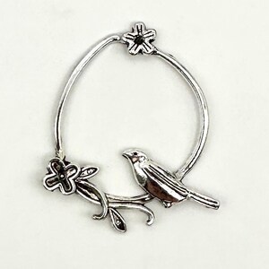 May include: Silver charm featuring a bird perched on a branch with a flower and a star. The charm is in the shape of a circle.