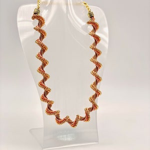 May include: A gold and orange beaded necklace with a spiral design. The necklace is displayed on a white mannequin. The text "Faux Cellini" and "Arcabeadies Designs" is visible at the top of the image.
