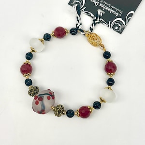 May include: A beaded bracelet featuring a mix of red, black, white, and gold-colored beads. The bracelet includes faceted red beads, black beads, and white oval beads. The clasp is gold-colored. The tag reads "Arcabeadies Design Handmade Jewelry & Accessories."