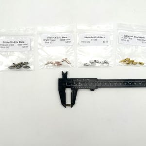 May include: Four bags of slide-on end bars in different finishes: antiqued brass, bright copper, steel, and gold tone. Each bag contains six bars that are 10mm long. A caliper is measuring one of the bars.