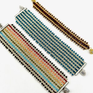 Könnte beinhalten: Drei Perlenarmbänder mit silbernen Verschlüssen. Die Armbänder sind aus kleinen, bunten Perlen in verschiedenen Mustern gefertigt. Das obere Armband ist braun und gold, das mittlere Armband ist türkis und silber, und das untere Armband ist ein Regenbogen aus Farben.