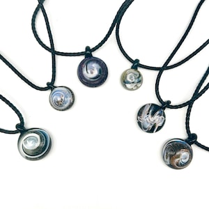 May include: A collection of handcrafted glass pendant necklaces. Each pendant features a unique swirling design in shades of blue, white, and black, suspended on a black cord. The pendants are round and vary slightly in size and pattern.