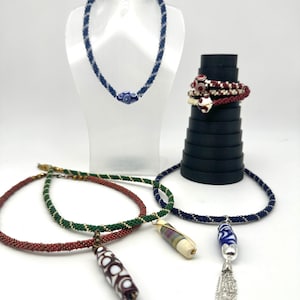 May include: A collection of beaded jewellery including a blue and white spiral necklace on a stand, a red and white beaded bracelet, and three necklaces with long cylindrical pendants in green, red, and blue with metallic accents and tassels.