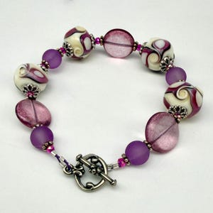 May include: A bracelet featuring round white beads with purple swirls, translucent purple disc beads, and matte purple spherical beads. The beads are accented with small silver-toned metal spacers and tiny pink crystals, secured by a silver-toned toggle clasp.