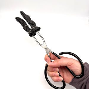 May include: A pair of black and silver tongs with black handles and fabric-covered tips. The tongs are held by a person against a white background. The tongs are designed for handling hot items.