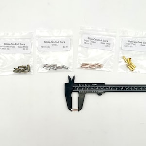 May include: Four bags of slide-on end bars in different finishes: antiqued brass, steel, bright copper, and gold tone. Each bag contains six 13mm bars. A caliper is measuring one of the copper bars.
