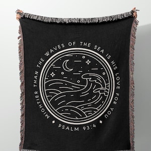 Op de afbeelding: Zwarte geweven sprei met witte tekst en een cirkelvormig ontwerp met golven, een maan en sterren. De tekst luidt "Mightier than the waves of the sea is his love for you" en "Psalm 93:4". De sprei heeft gefranjerde randen.