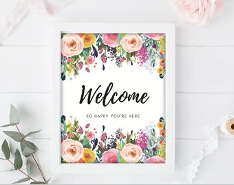 Könnte beinhalten: Ein weiß gerahmtes Schild mit dem Wort "Welcome" in schwarzer Schrift, umgeben von einem floralen Aquarellrahmen aus rosa, gelben und lila Blumen. Der Text "So happy you're here" steht darunter.