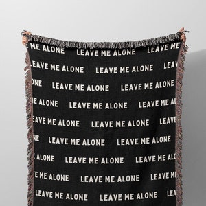 May include: Black throw blanket with white text that reads "LEAVE ME ALONE" repeated across the surface. The blanket has a fringed edge and is displayed against a neutral background.