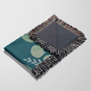 Op de afbeelding: Teal plaid deken met een botanisch bladerpatroon in lichtgroen en wit. De deken heeft een gefranjerde rand met een mix van kleuren. De deken is opgevouwen en toont het patroon en de achterkant.